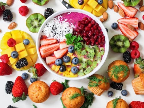 Colorful smoothie bowl topped with dragon fruit, pomegranate, kiwi and berries, surrounded by fresh fruit and muffins