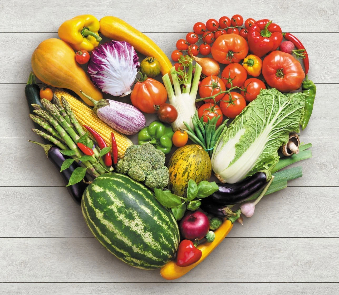 Fresh vegetables and fruit including tomatoes, peppers, corn and broccoli arranged in a heart shape on a wooden surface