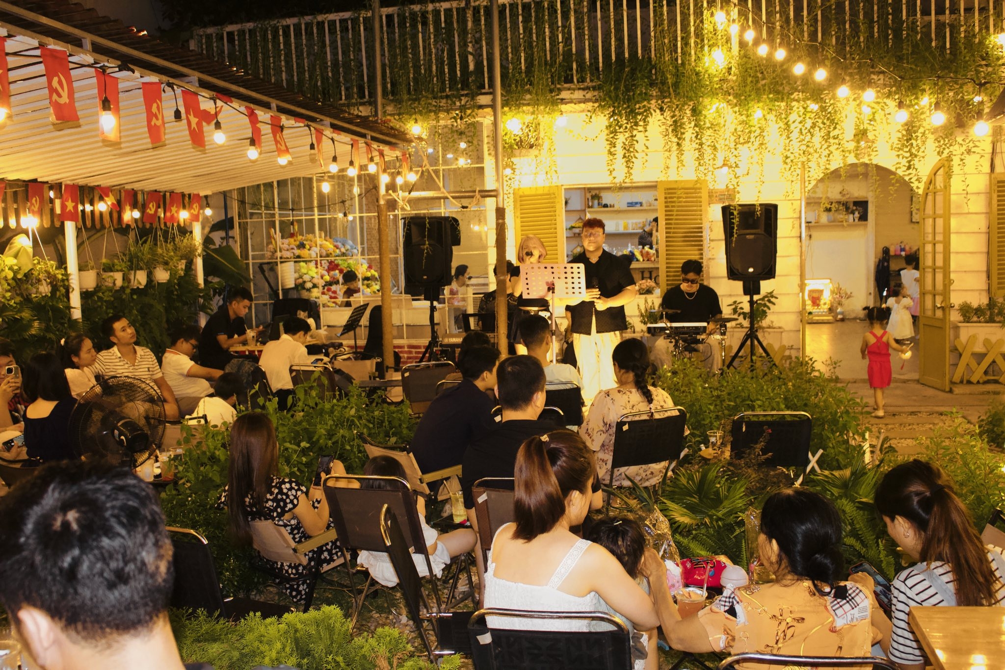 Outdoor garden cafe at night with warm string lights and flags, an audience seated as a musician performs on a small stage