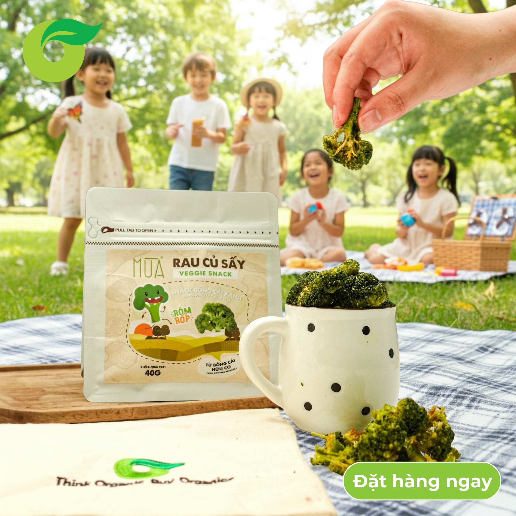 A pack of dried broccoli veggie snacks and a polka-dot mug at a sunny park picnic with children playing in the background