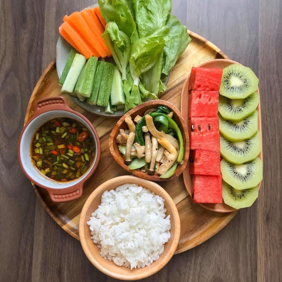 A wooden tray with fresh carrot and cucumber sticks, lettuce, mushrooms, white rice, dipping sauce, watermelon and kiwi slices
