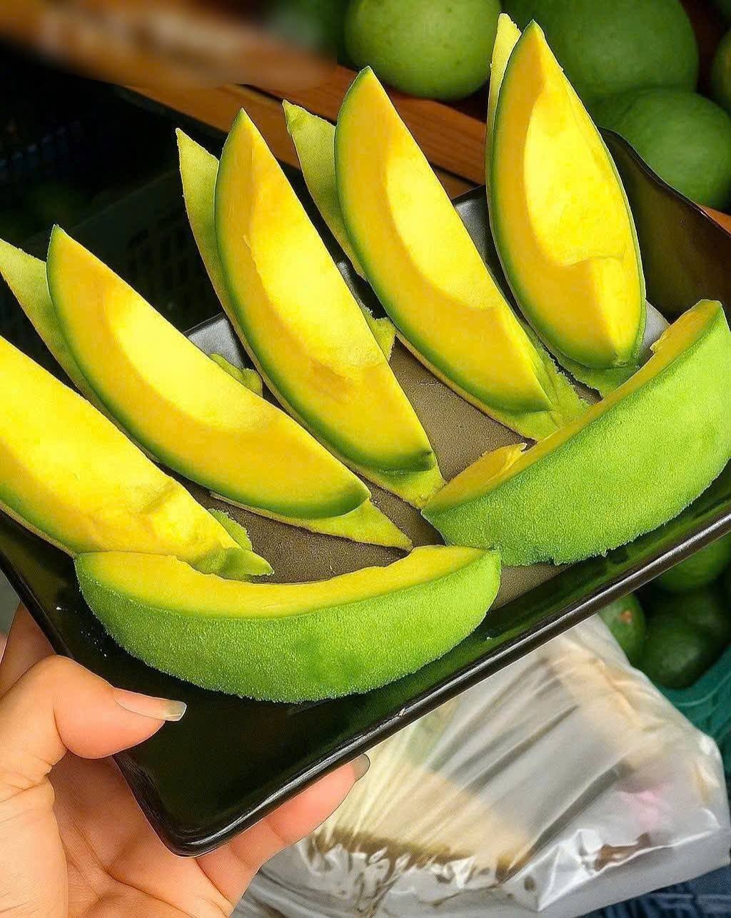 A hand holding a black tray of sliced green mango wedges with bins of whole green mangoes in the background