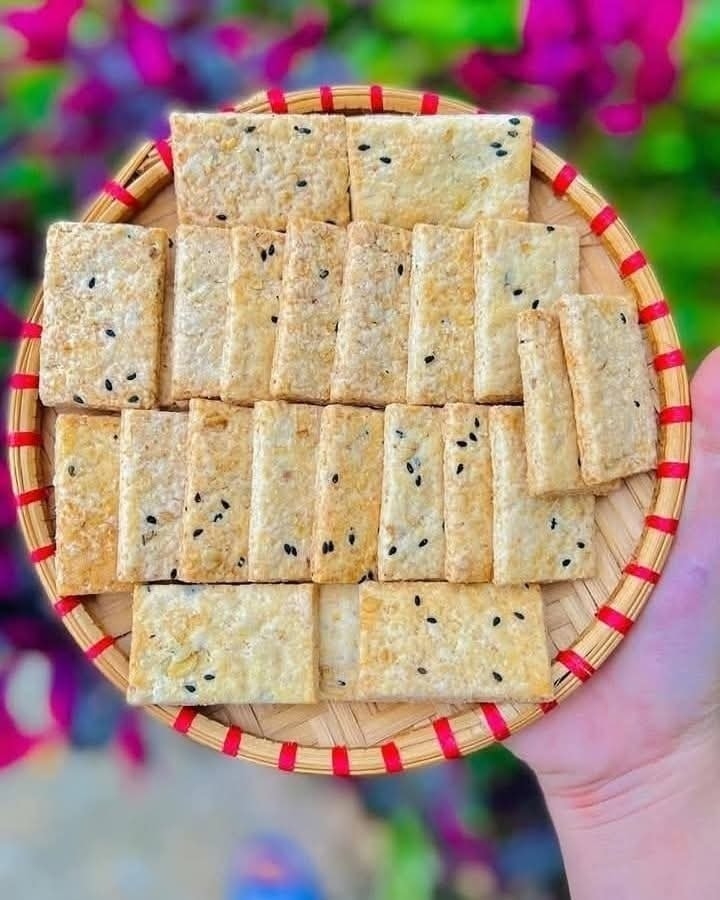 A hand holding a round bamboo tray of crispy black-sesame rice crackers against a colorful bougainvillea background