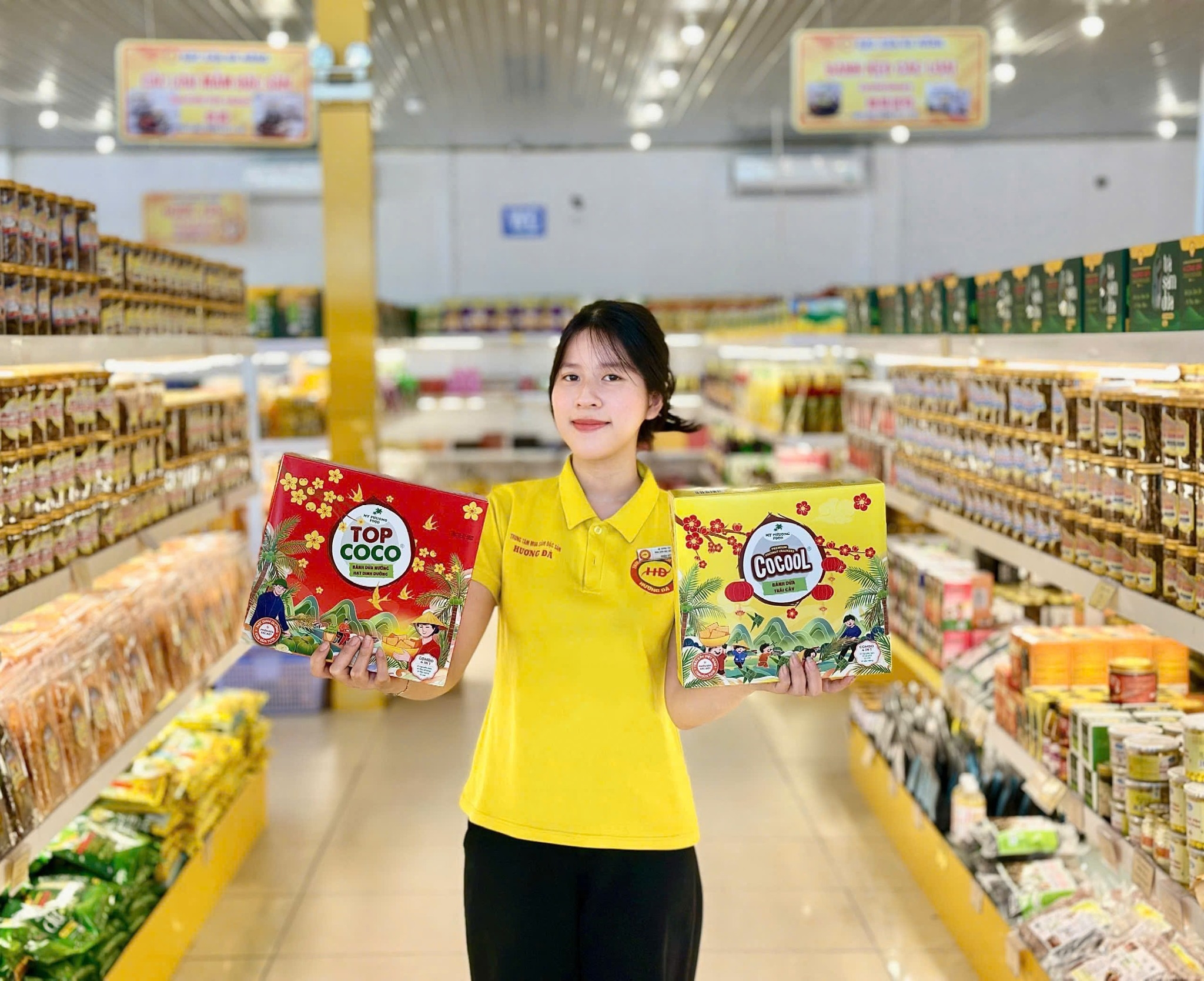 Staff member in a yellow shirt holding two colorful gift boxes in a specialty store aisle stocked with products