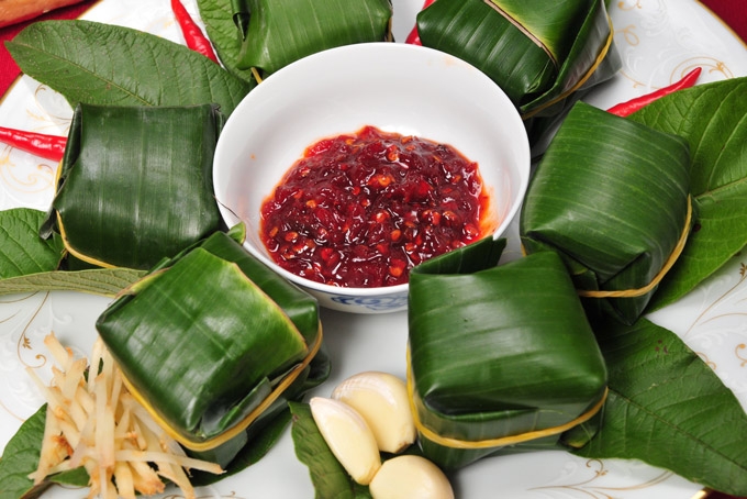 Banana leaf wrapped fermented pork rolls arranged around a white bowl of red chili dipping sauce with garlic