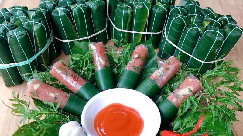 Banana leaf wrapped fermented pork rolls with herbs stacked behind a white bowl of red dipping sauce