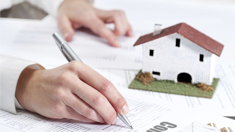 Hand signing a property contract with a pen beside a small white model house with a red roof
