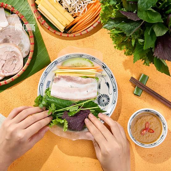 Hands rolling soft rice paper around sliced pork and fresh herbs at a Da Nang table, with a bowl of mắm nêm alongside