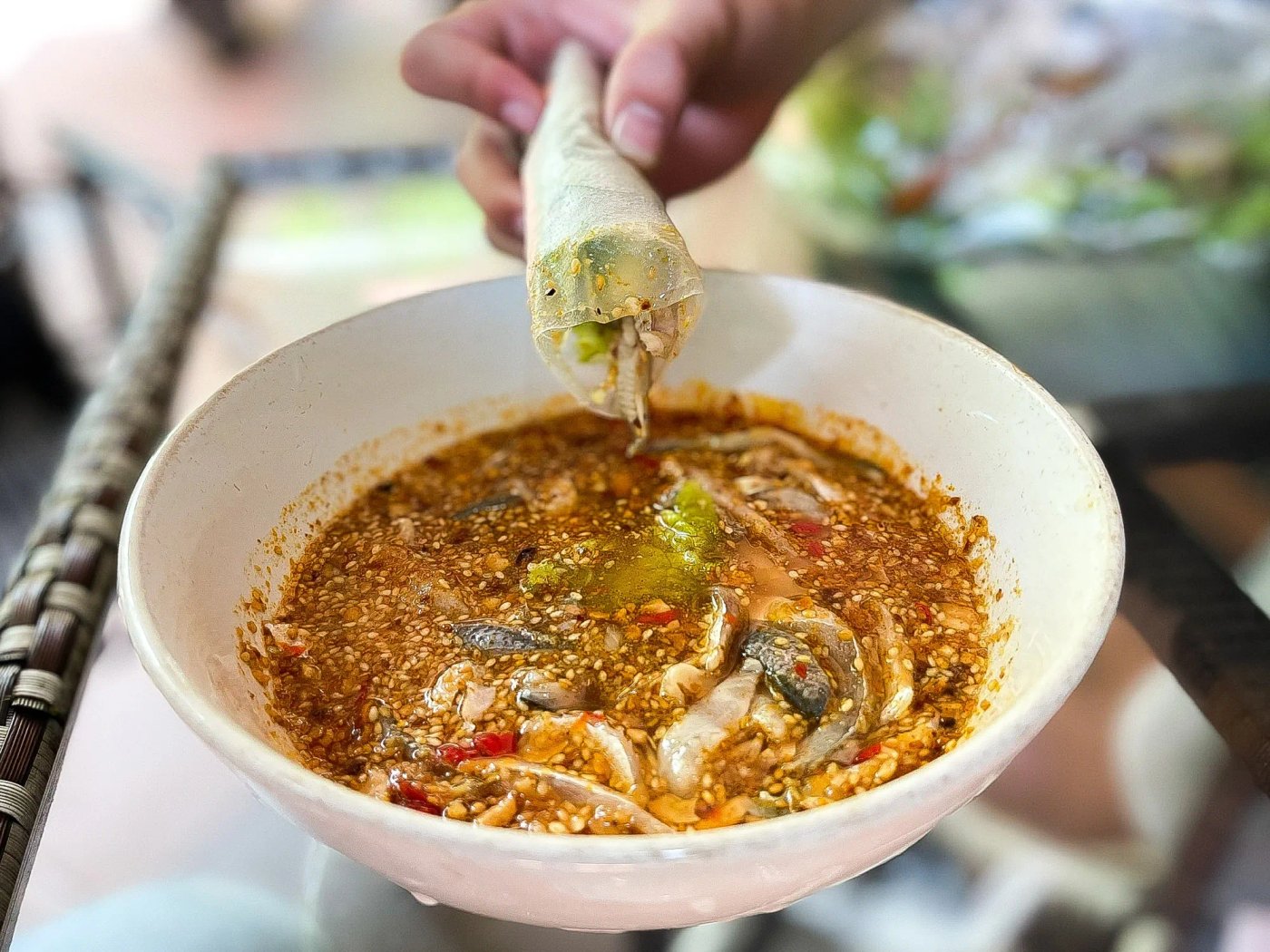 A rice-paper roll of gỏi cá being dipped into a bowl of thick sesame-peanut-chili sauce