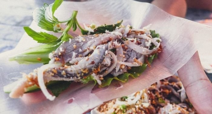 A hand holding a gỏi cá Nam Ô wrap — fish, shredded onion and herbs on a leaf and rice paper, ready to eat