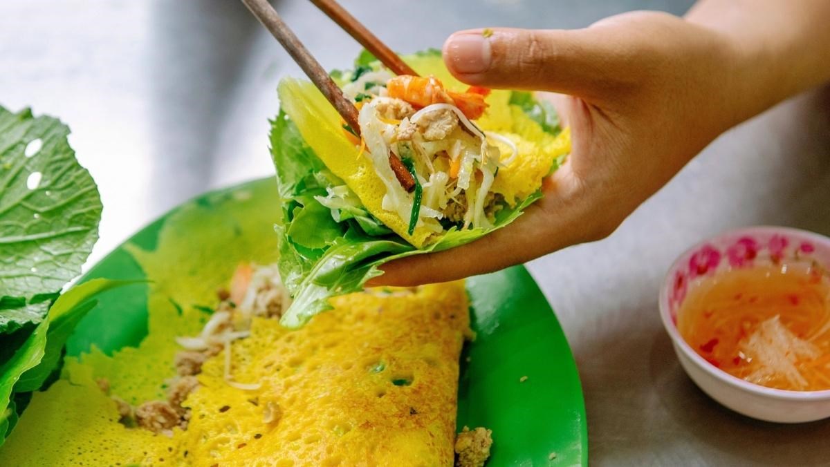 A hand holding a piece of bánh xèo wrapped in lettuce and herbs, dipping it into a bowl of nước chấm