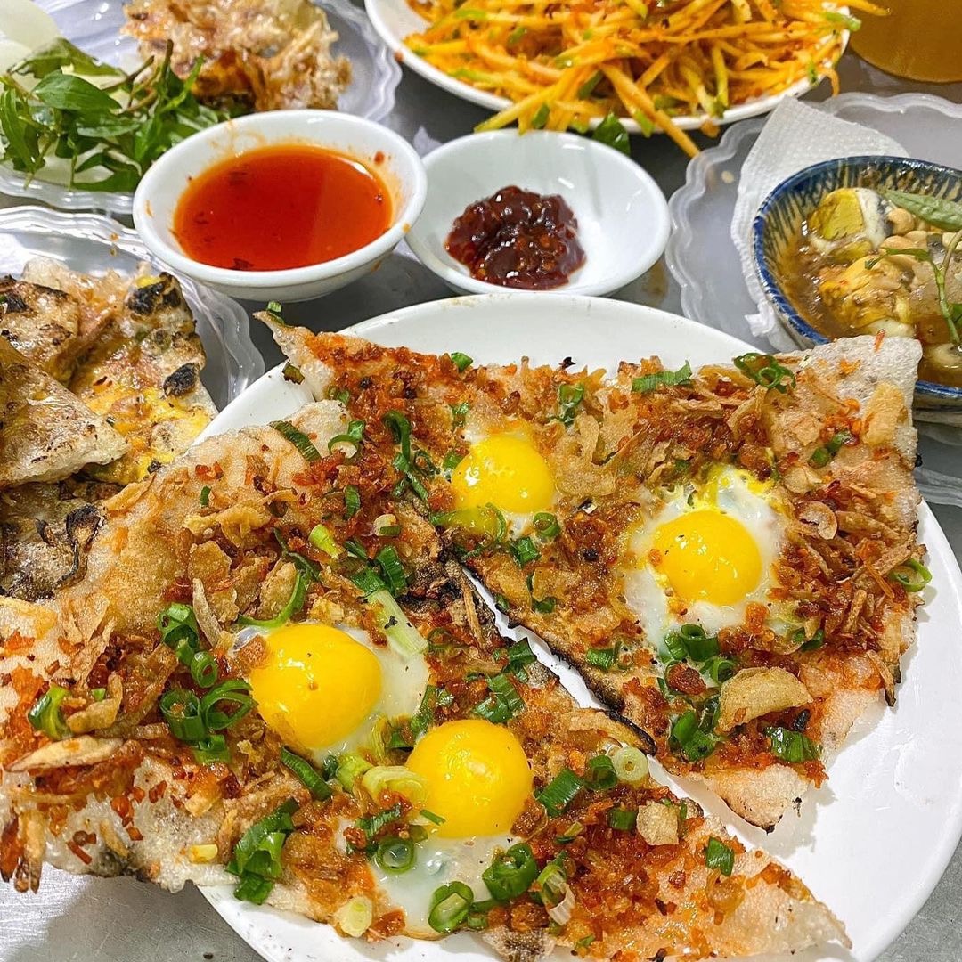 Close-up of bánh tráng kẹp — crispy grilled rice paper topped with quail egg, dried floss and scallion, with dipping sauces alongside.