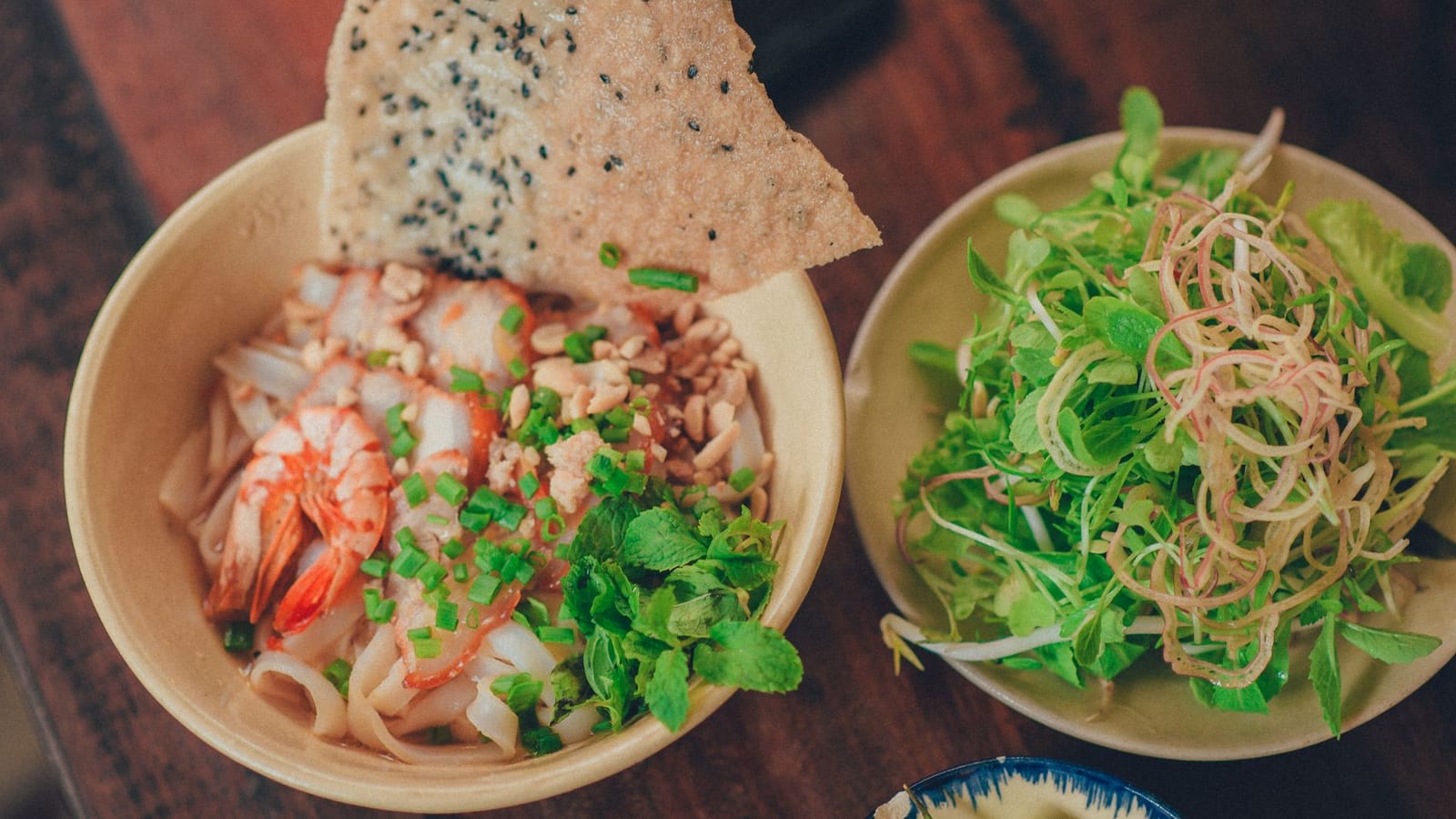 A close-up bowl of mì Quảng with wide yellow noodles, shrimp, pork, peanuts and fresh herbs