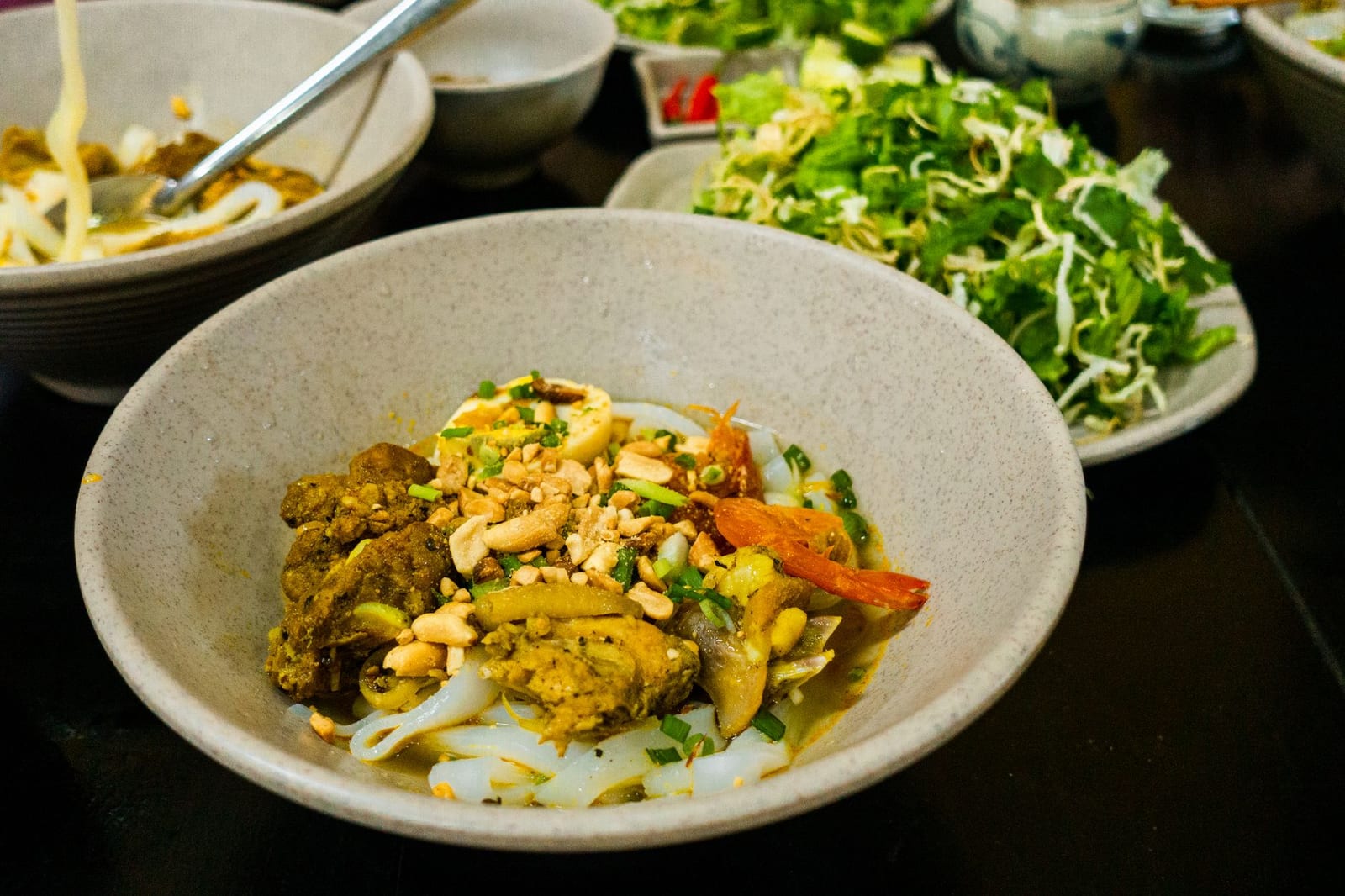 A bowl of mì Quảng with wide turmeric-yellow rice noodles, pork, shrimp, peanuts and herbs, with a side plate of greens