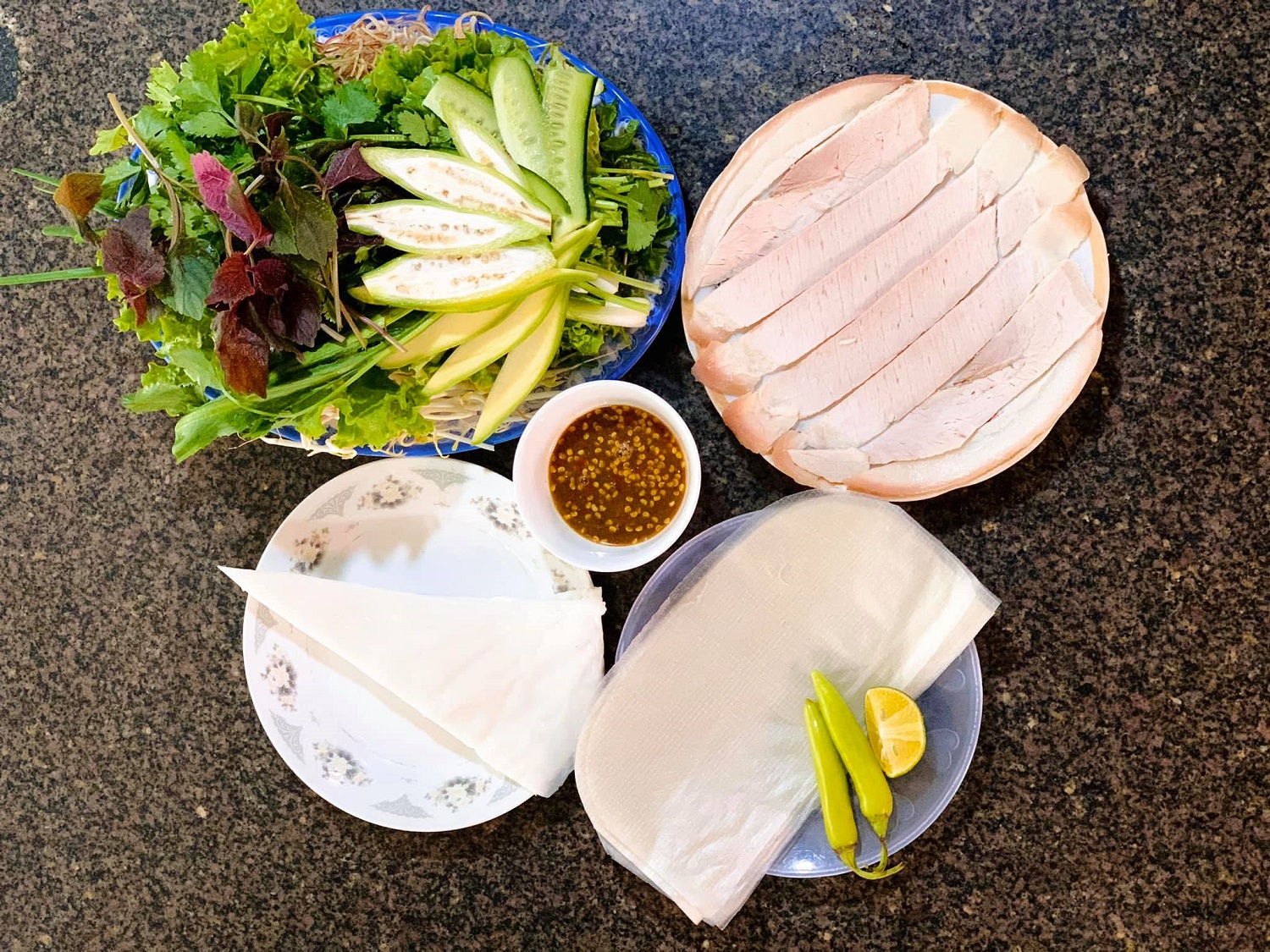 A bánh tráng cuốn thịt heo spread — sliced boiled pork, a basket of fresh herbs and green banana, rice paper, and a bowl of mắm nêm with chili and lime