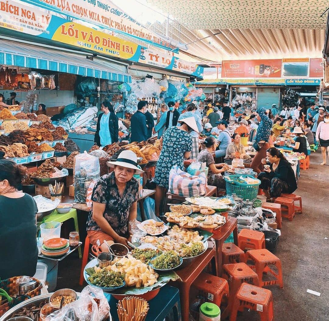 A busy food-court aisle inside a Da Nang market, lined with vendors, plastic stools and plates of local street food