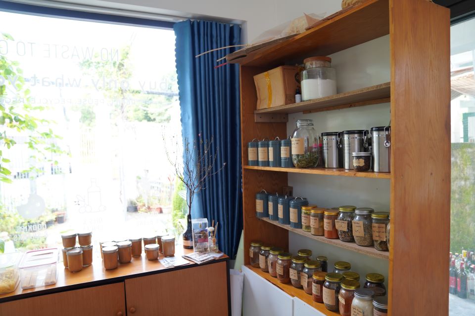 Wooden shelving wall at No Waste To Go lined with glass jars of bulk dry goods, with a window looking onto a green courtyard.