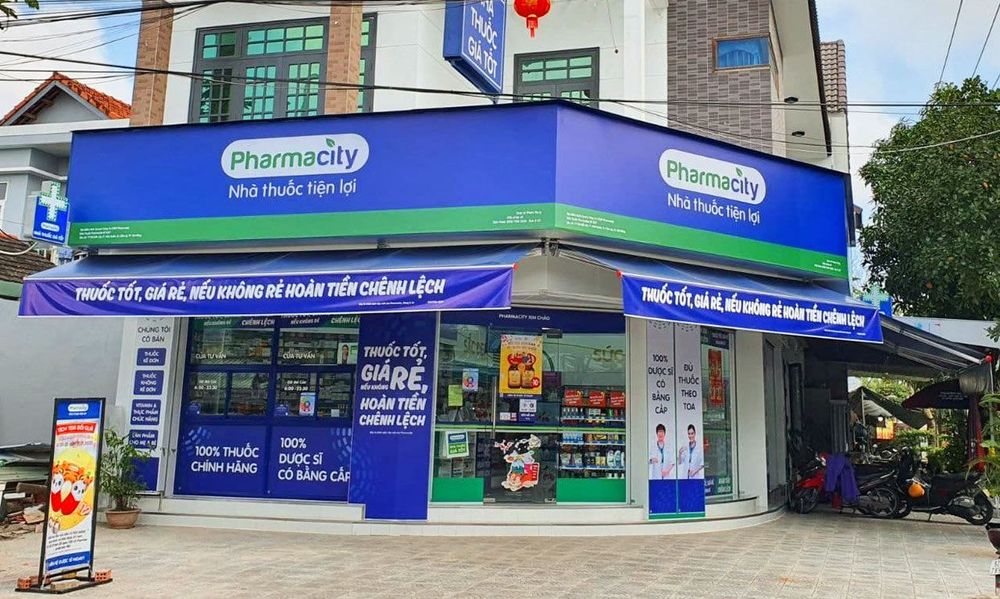 A Pharmacity branch on a Vietnamese street corner, its blue and green shopfront advertising '100% thuốc chính hãng' (100% genuine medicine) and '100% dược sĩ có bằng cấp' (100% qualified pharmacists) — the visual shorthand of a modern chain pharmacy